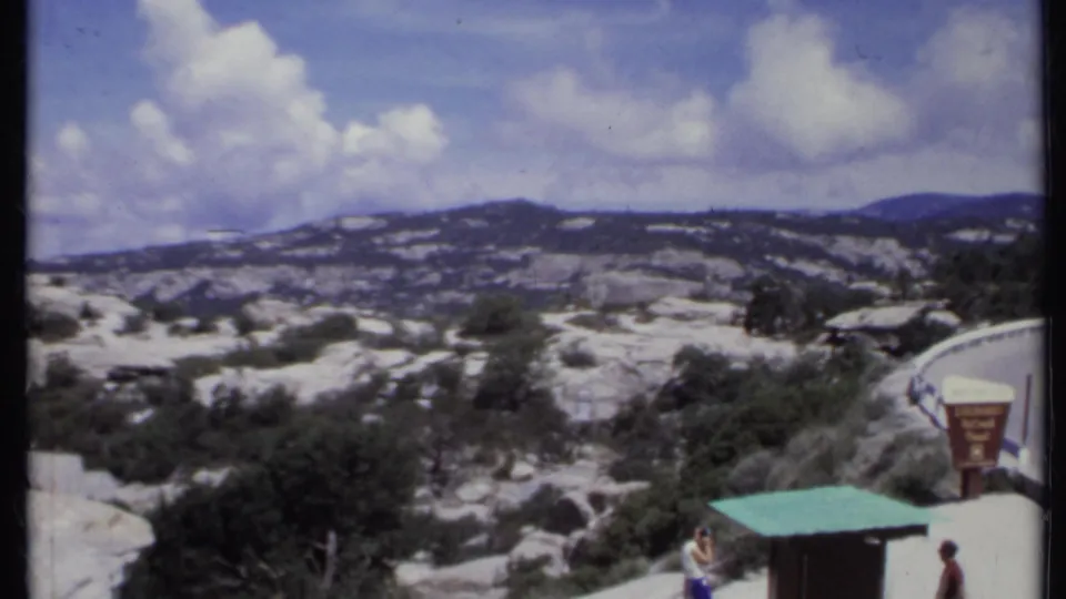 1971 California 8mm view across rocky hills from a roadside overlook, with visitors and a small green-roofed structure near the road.