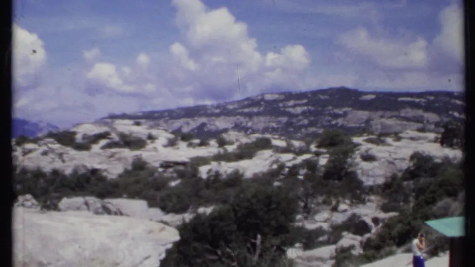 1971 California home-movie frame holding on a rocky mountain vista with scattered greenery, distant ridges, and a bright clouded sky.