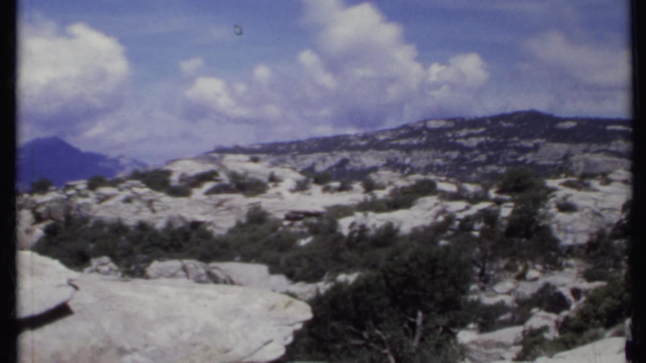 Rocky mountain overlook with shrubs, distant ridges, and clouded blue sky.