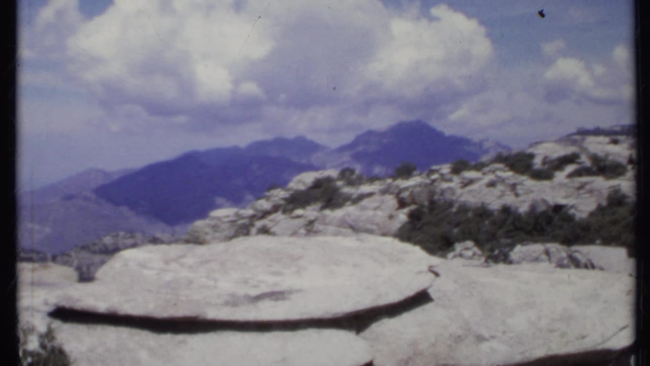 Flat foreground rock with mountain ridges and shrubs beneath large clouds.