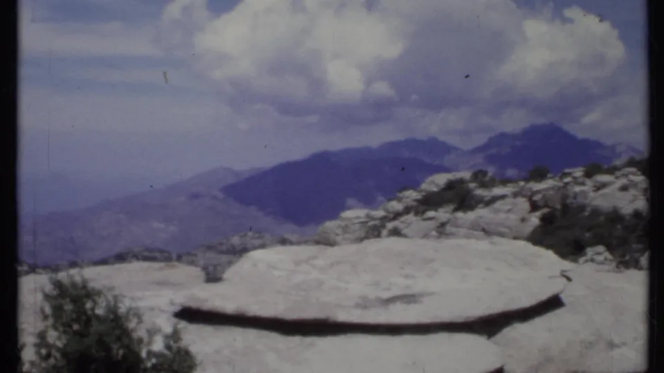 1971 California 8mm frame with a broad pale rock ledge, overlook railing, shrubs, and layered mountain ridges.