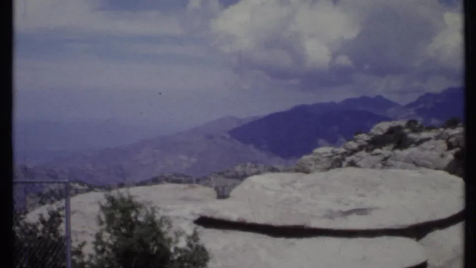 1971 California landscape frame showing rocky foreground, sparse shrubs, a railing, hazy valley, and distant mountains.