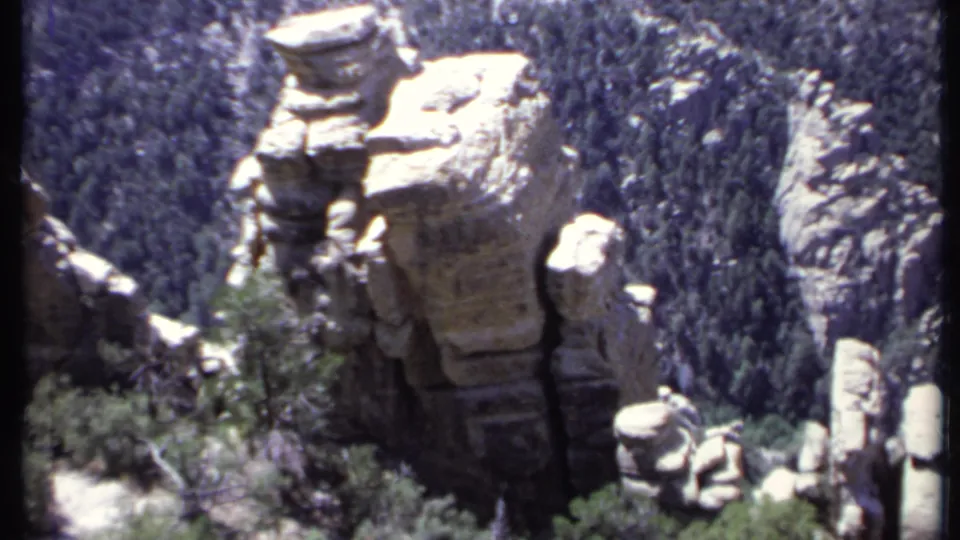 1971 California 8mm frame showing a large weathered rock tower, nearby shrubs, and dark evergreen forest behind it.