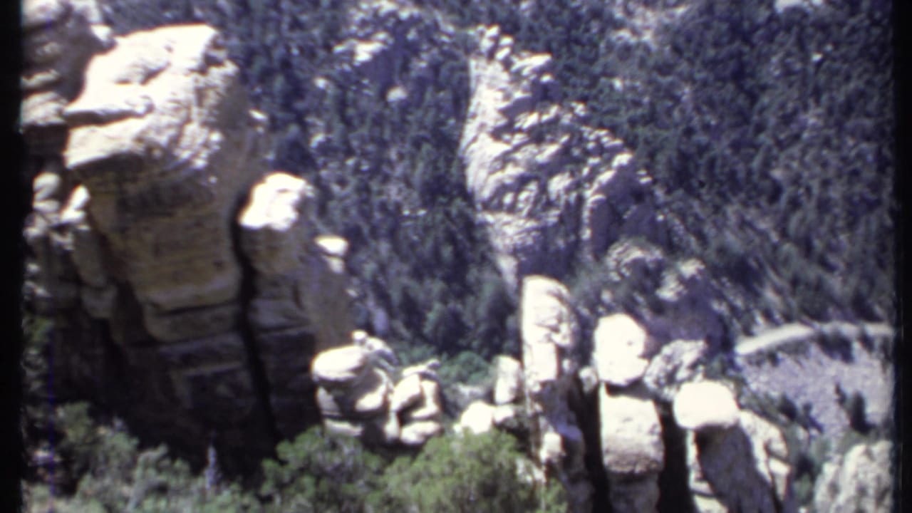 1971 8mm California mountain view with pale rock formations, evergreen-covered slopes, and a winding road or trail below.