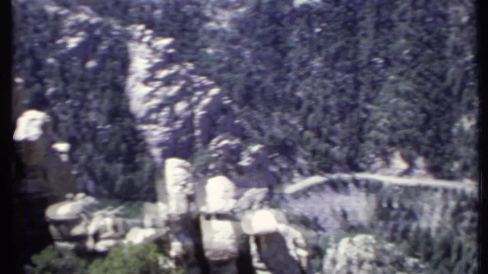 8mm-era California mountain landscape showing rock columns, dense evergreens, and a narrow road or trail crossing the slope.