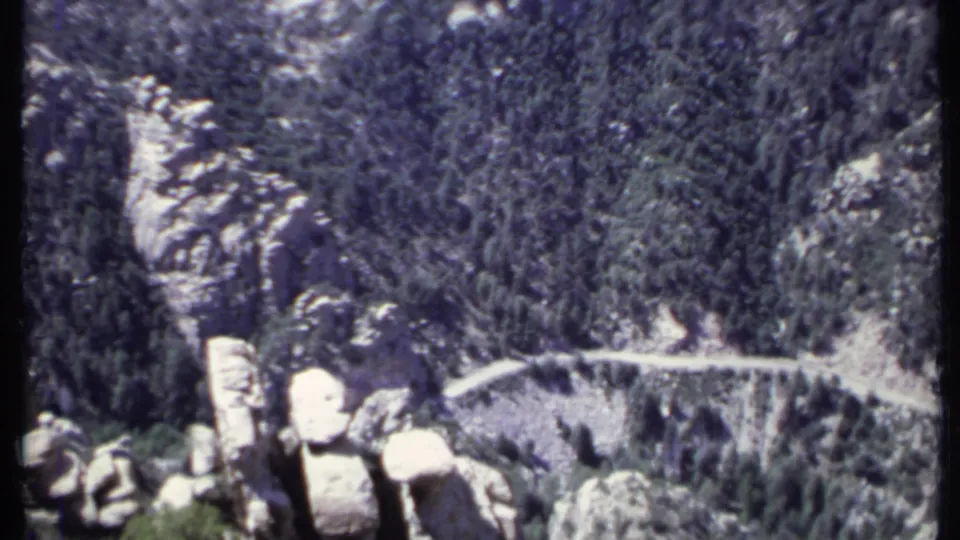 1971 home-movie frame of a California mountain slope with evergreen forest, pale cliffs, and a winding road or trail.