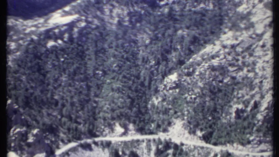 Soft 8mm frame of California mountain terrain, with evergreen-covered slopes and a road or trail bending through the rocky valley.