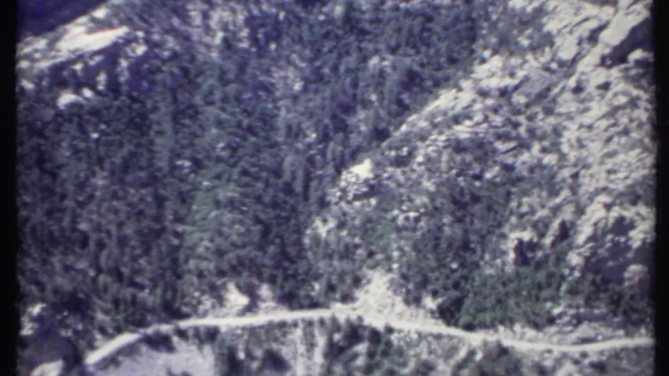 1971 California 8mm frame showing a narrow dirt road cut across a steep evergreen and rocky mountainside.