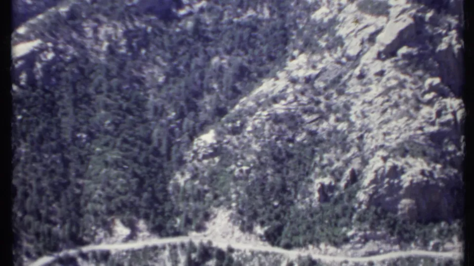 1971 California home-movie frame of a winding unpaved road beneath dense trees and exposed rock.