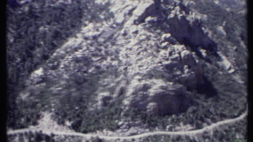 1971 California home-movie frame showing a curving dirt road below a steep rocky and wooded mountainside.