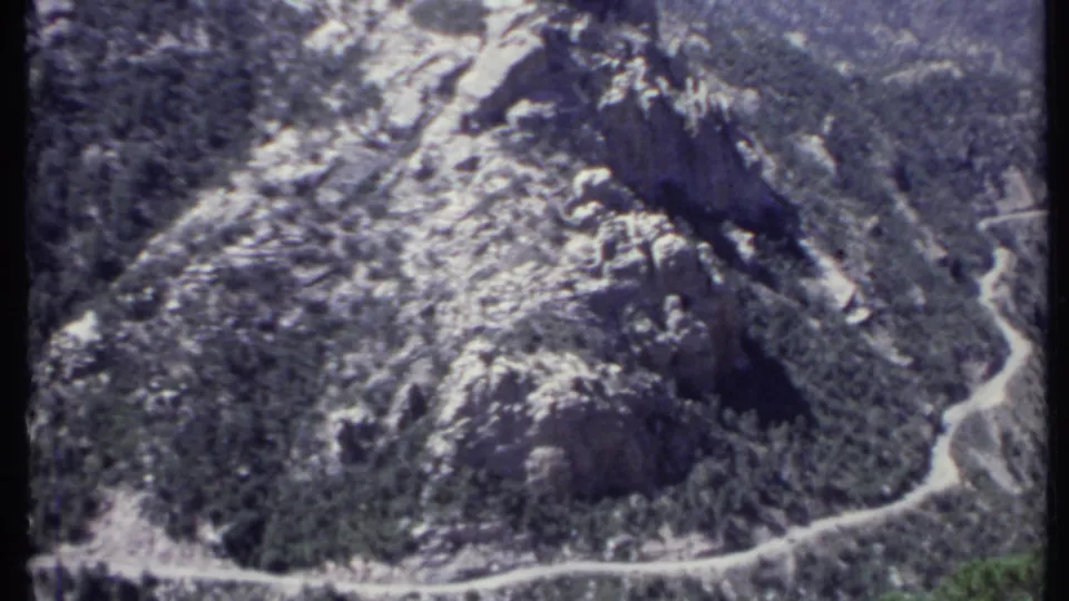 1971 California 8mm aerial frame showing rugged mountain slopes, patchy snow, and a road curving through the valley below.
