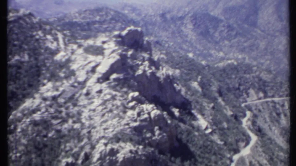 Aerial 8mm view over California mountain ridges with light snow, dark forest patches, and hazy terrain in the distance.