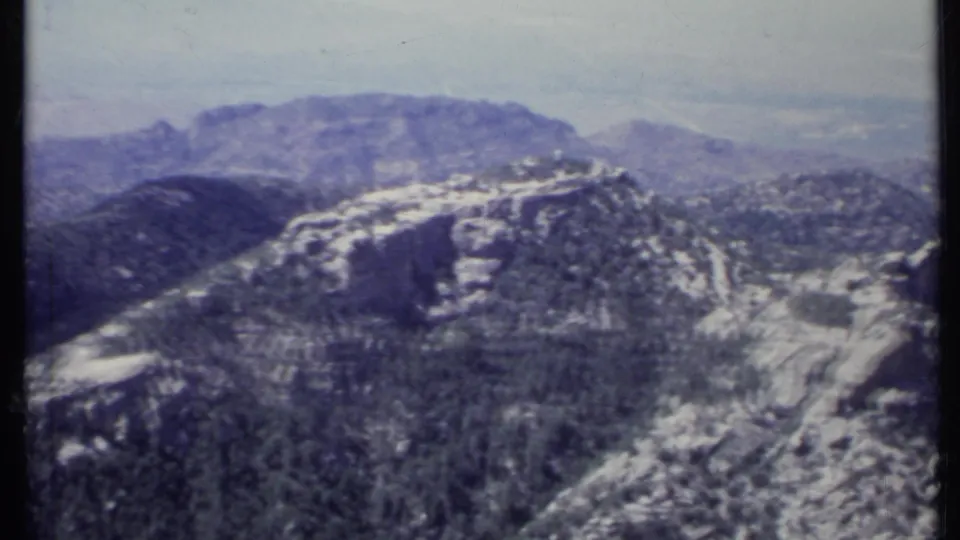 Aerial 8mm frame of California mountain terrain with snow-streaked rock, forested slopes, and hazy ridgelines beyond.