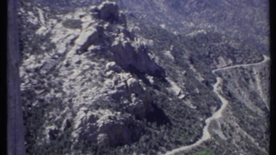 1971 California 8mm frame showing a rugged rocky mountain slope with scattered trees and a winding road below.