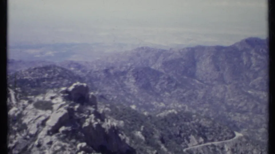 1971 California 8mm frame of a broad mountain valley, rocky foreground ridge, sparse trees, and hazy distant ranges.