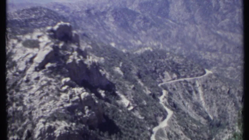 1971 California 8mm view of a rocky mountain outcrop above wooded hills, with a pale winding road cutting across the slope.