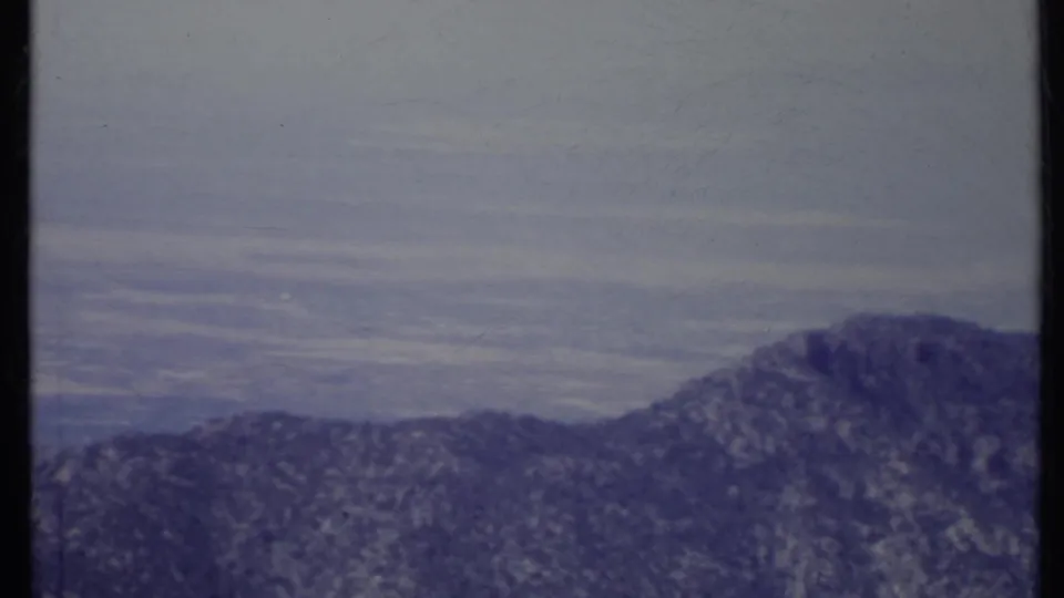 1971 California 8mm frame showing a dark mountain slope, distant hazy valley, and subdued overcast sky.
