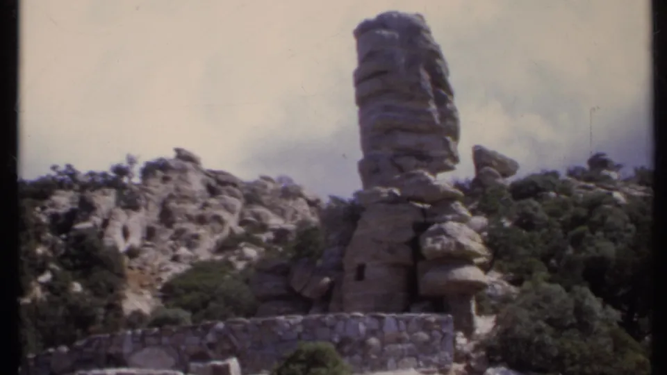 1971 California 8mm frame showing a tall natural rock spire or stacked formation with shrubs, boulders, and a low stone wall.