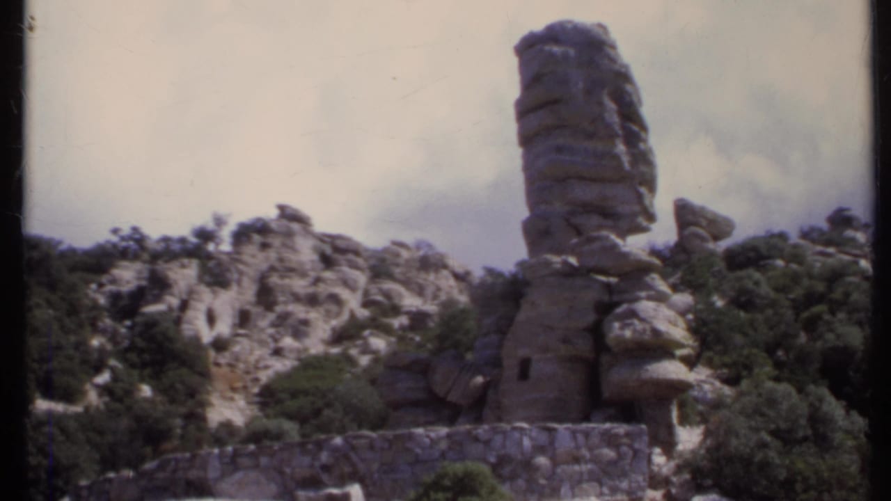 Layered stone column and rocky hillside above trees under a cloudy sky.