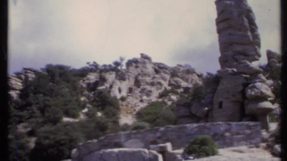 A visitor-shot 8mm view of a vertical rock formation, stone wall, shrubs, and weathered cliffs beneath a clouded sky.