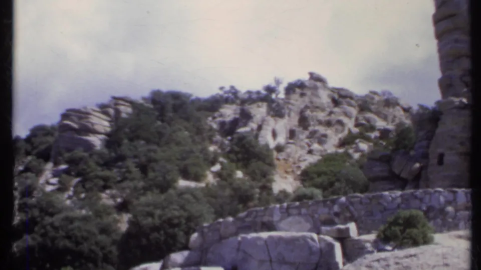 Soft 8mm frame showing scrub trees, a stone wall, rocky slope, and the edge of a tall layered formation in 1970s travel footage.