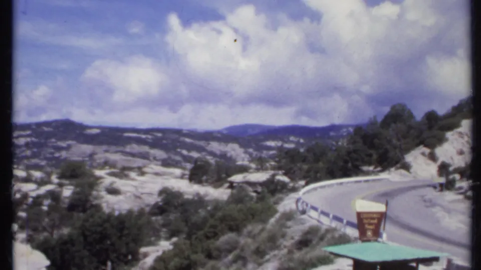 1971 8mm roadside view of a winding road, guardrail, hills, trees, and a visible Colorado National Monument sign.