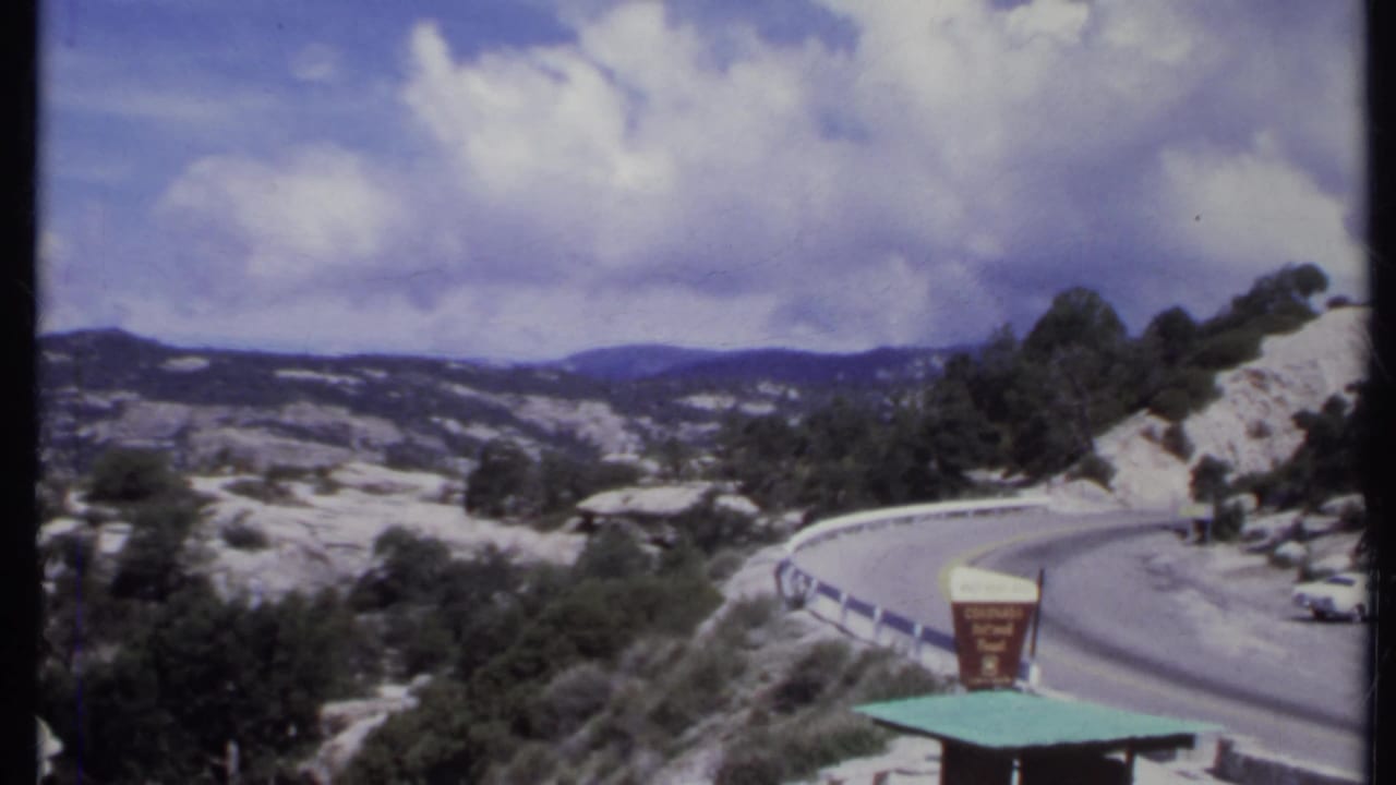 Curving mountain road with rocky hills, guardrail, sign, and cloudy sky.