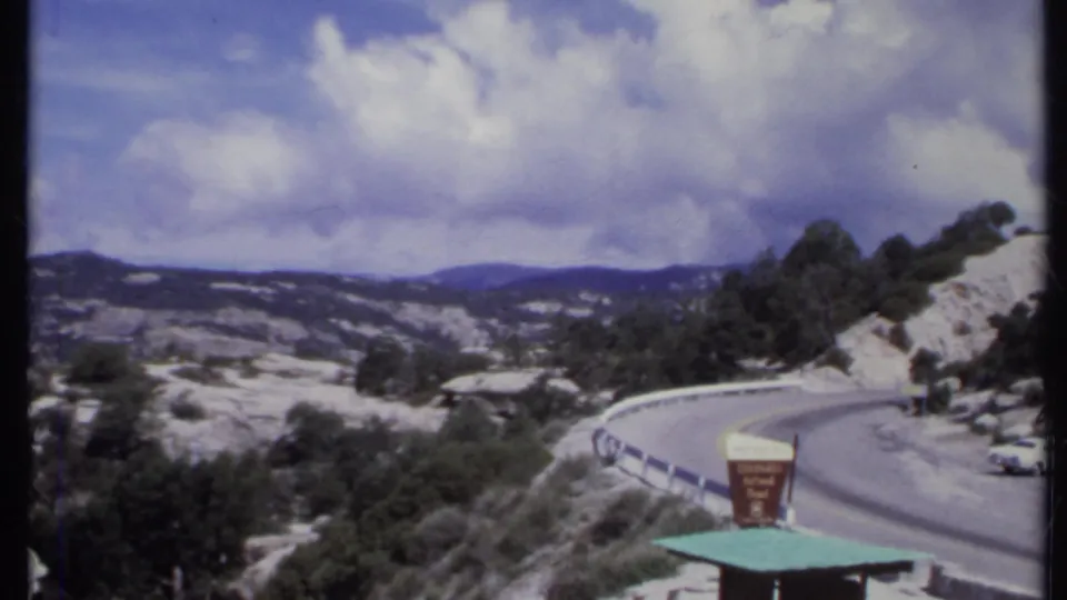 1971 California 8mm frame showing a curving mountain road through rocky terrain with a guardrail, roadside sign, and cloudy sky.