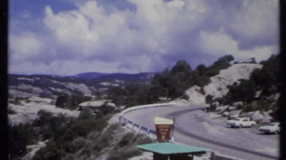 1971 California 8mm frame of a scenic overlook beside a winding mountain road, with rocky slopes, a sign, and white parked cars.