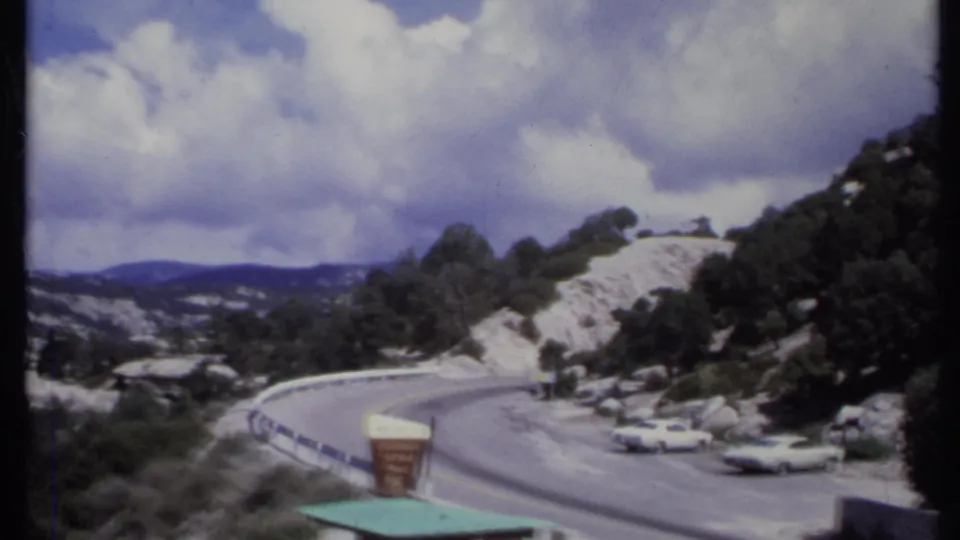 1971 California 8mm frame showing a winding road at a rocky scenic pullout, with a roadside sign, parked white cars, and cloud cover.