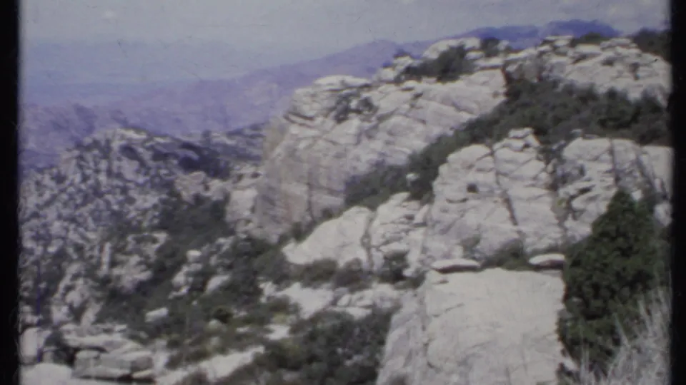 1971 California 8mm frame looking across pale rock formations, shrubs, and a hazy mountain valley from a scenic overlook.