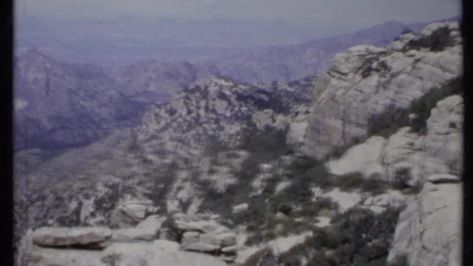 1971 California 8mm frame with jagged rock formations, sparse shrubs, layered ridges, and distant hazy terrain.