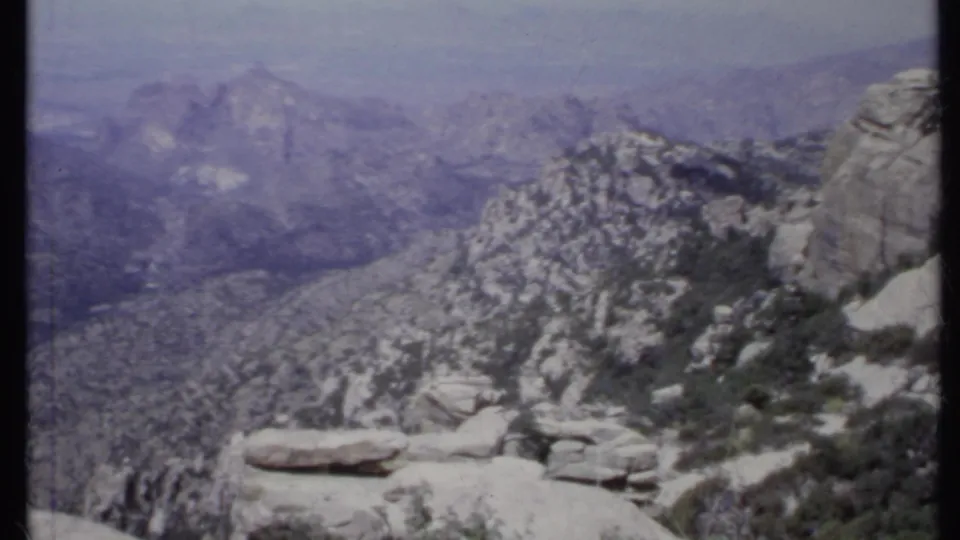 1971 California 8mm frame showing pale rock ledges, scrubby plants, steep mountain terrain, and layered ridges in haze.