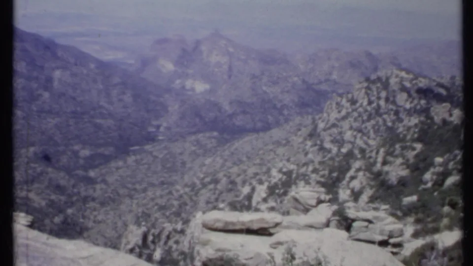 1971 California 8mm frame with a wide view over rugged ridges, pale rocks, sparse foliage, and distant hazy slopes.