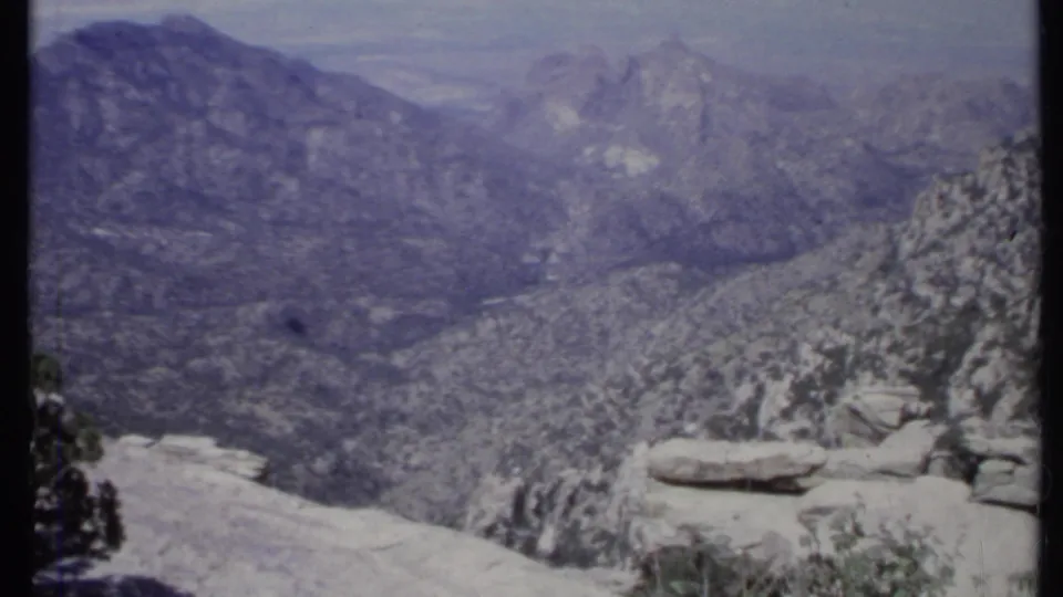 1971 California home-movie frame with distant ridges, foreground boulders, scrub vegetation, and a hazy blue-purple cast.