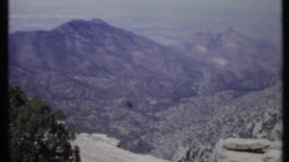 1971 California 8mm frame showing a hazy mountain ridge, valley terrain, pale rock, and dark trees near the lower foreground.