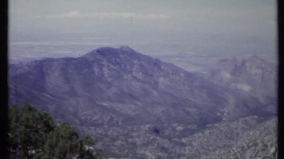 1971 California 8mm frame with distant peaks, broad ridgelines, foreground trees, and rocky terrain softened by haze and film grain.