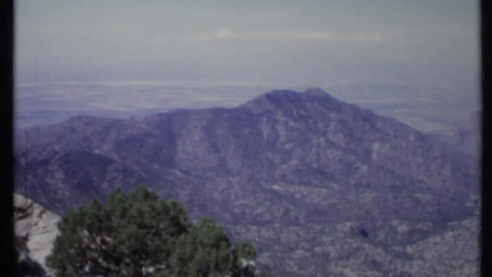 1971 California 8mm view of rugged mountains, foreground evergreens, and a broad hazy valley under a pale sky.