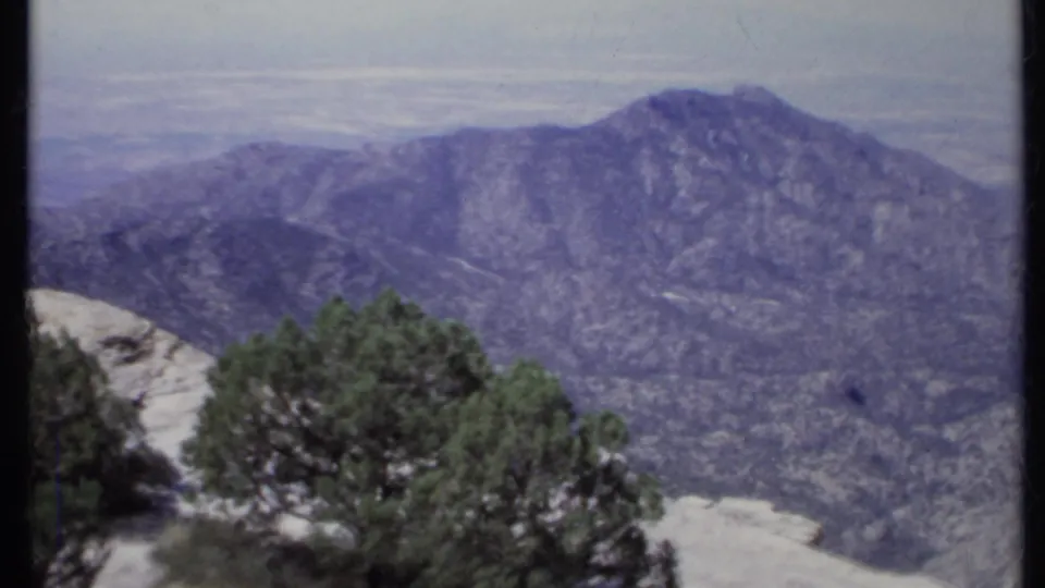 1971 California 8mm frame looking from a rocky overlook toward a mountain slope and hazy valley beyond.