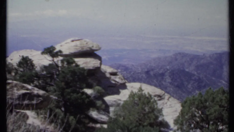 1971 California 8mm frame with a rocky overlook, dark evergreens, and blue-gray ridgelines fading into haze.