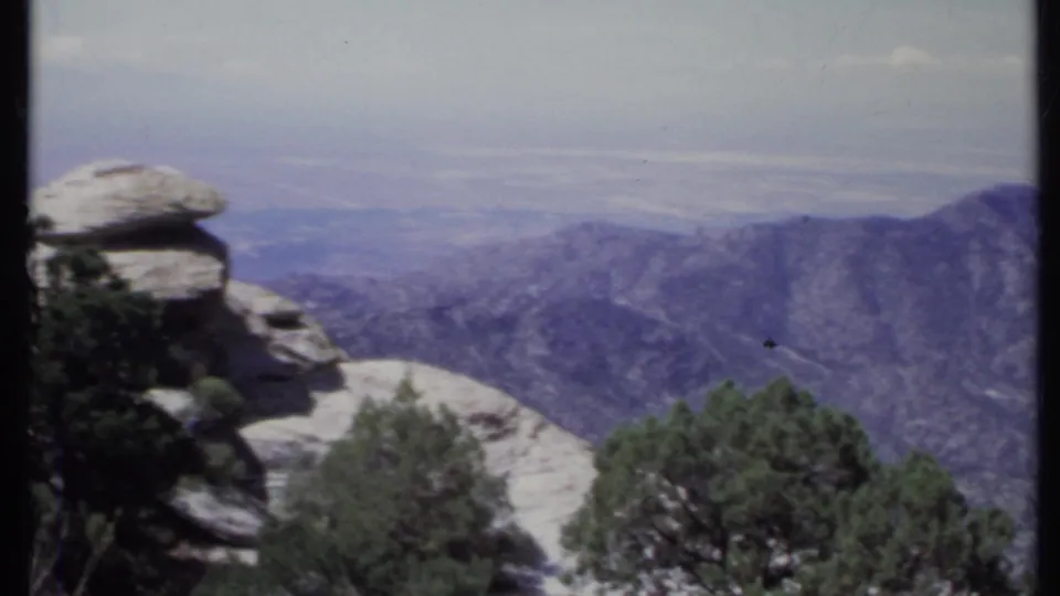 1971 California 8mm frame from a slow pan, showing a rock formation, foreground evergreens, and distant mountain terrain.