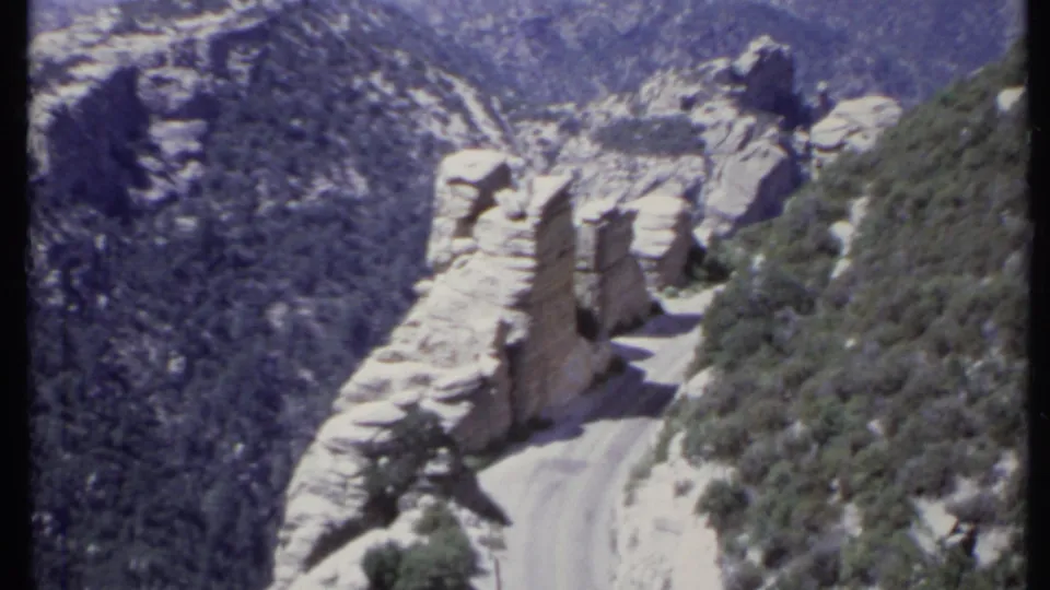 1971 California 8mm frame of a winding dirt road bordered by pale outcrops and scrub on a steep mountain pass.