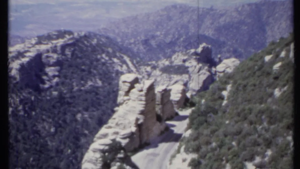 A 1971 California home-movie frame looking across a rocky mountain road toward hazy ridges and brush-covered slopes.