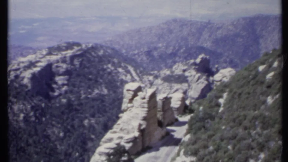 Final 1971 California 8mm frame showing the narrow cliffside road, pale stone, brushy slope, and distant mountain haze.