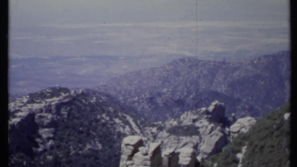 1971 California 8mm frame with a rugged foreground ridge, pale stone formations, wooded slopes, and a hazy valley in the distance.