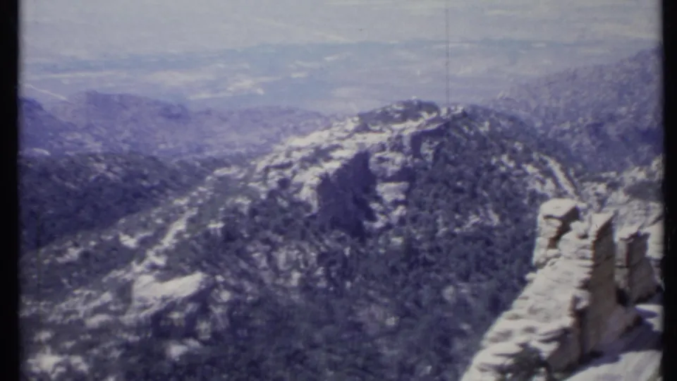 1971 California 8mm frame showing a rocky overlook edge, forested mountain slopes, layered ridges, and a hazy valley beyond.