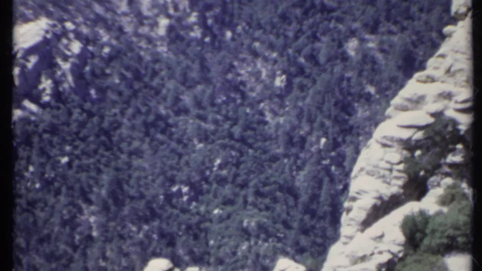 1971 California 8mm frame with a vertical rock wall and dark pine-covered hillside beyond it.