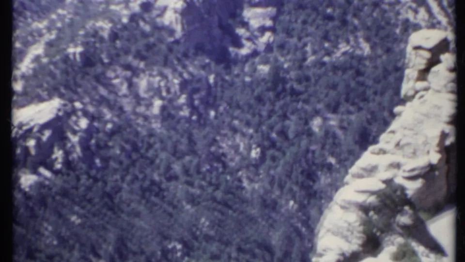 1971 California home-movie frame showing pine forest, rocky outcrops, and a pale cliff edge across a canyon.