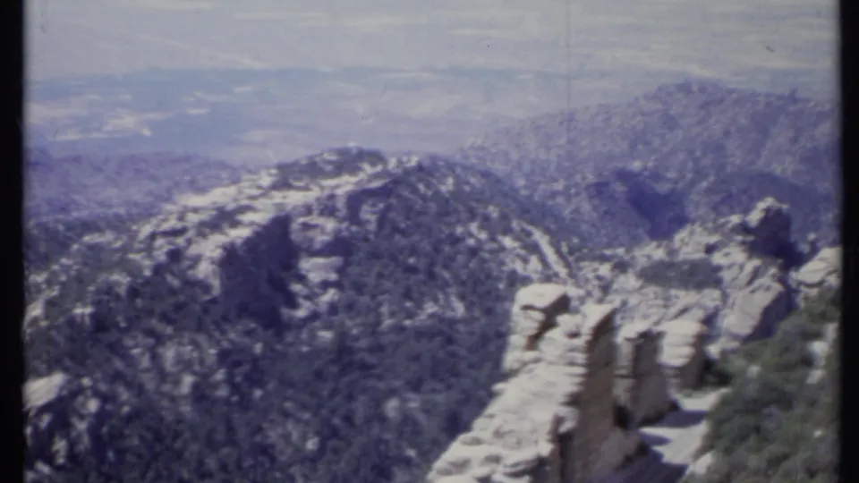 1971 California 8mm view of a rocky foreground ledge, wooded slopes, and layered mountain ridges fading into haze.