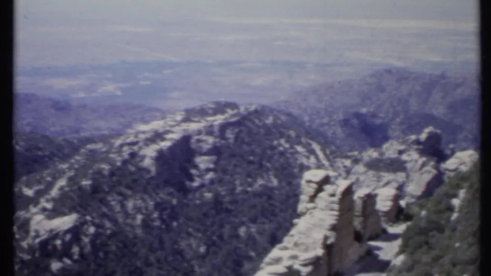 1971 California 8mm frame with snow-streaked ridge, tan rock formations, evergreen slopes, and hazy valley distance.